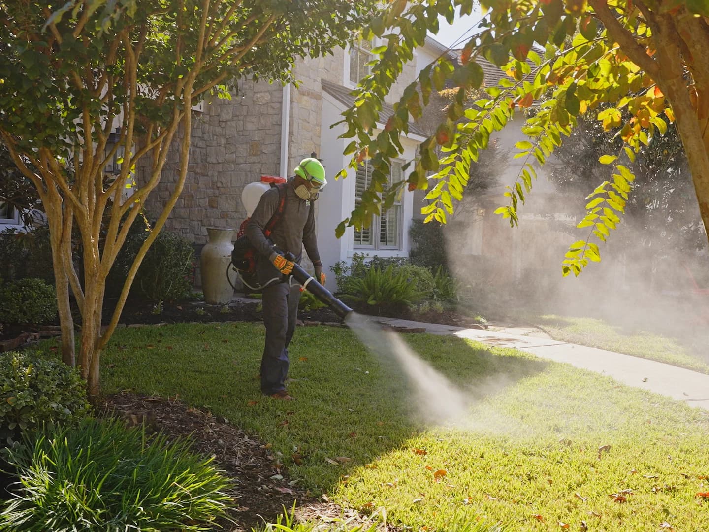 Technician in a respirator and high-visibility hard hat applies a mosquito barrier treatment with a backpack mister across a suburban front lawn.