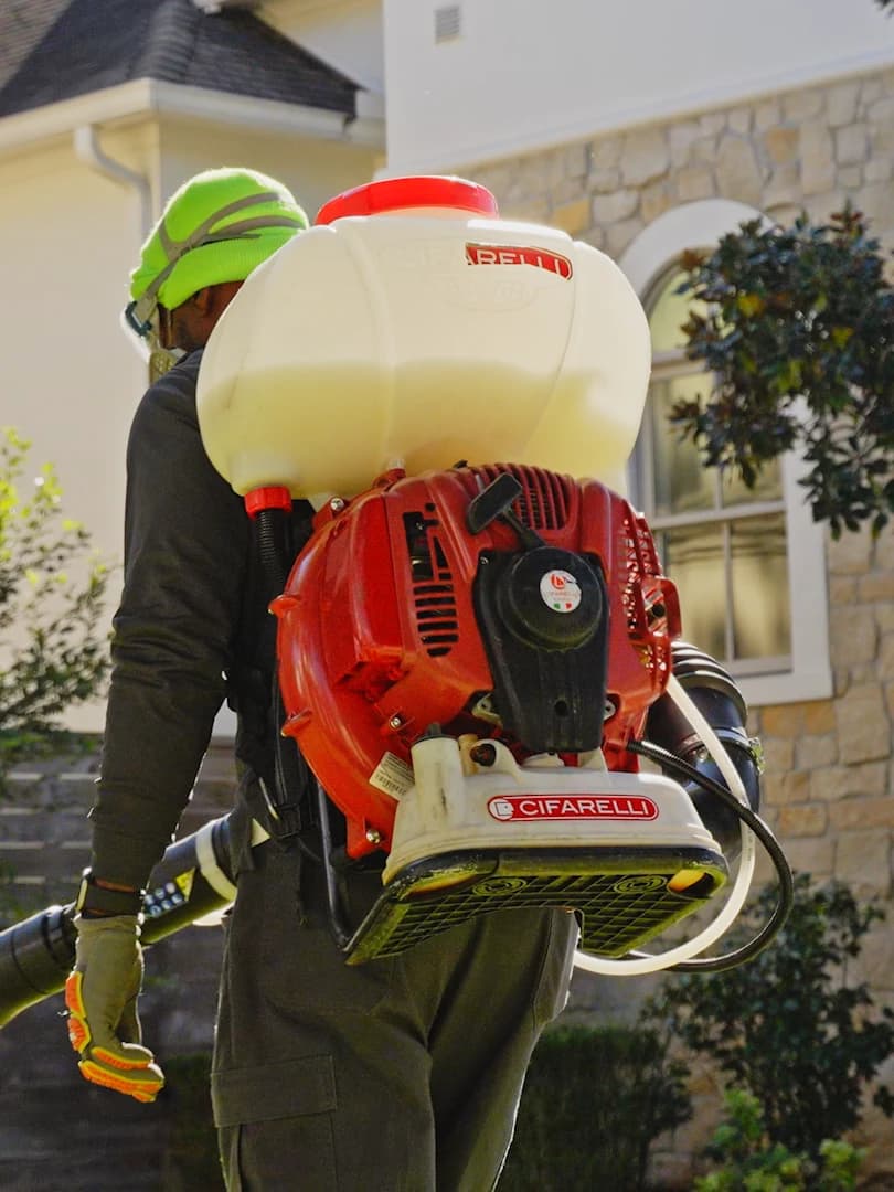 Mosquito control technician wearing a backpack mist blower in the yard of a stone-sided home, ready to apply a barrier treatment around the house and landscaping.